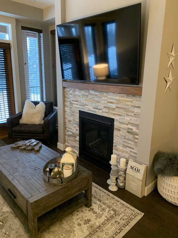Un salón con chimenea, un televisor de pantalla plana montado encima, una mesa de centro con adornos y una zona de estar confortable con sofá y sillón.