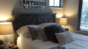 Ein Schlafzimmer mit einem Bett, zwei Nachttischlampen, einem Nachttisch, einem Fenster mit Jalousien und einem Wandschild mit der Aufschrift 'ANTIQUES FLEA MARKET'.