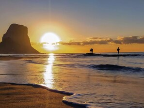 A beach scene with a large rock formation and the sun setting over the ocean.