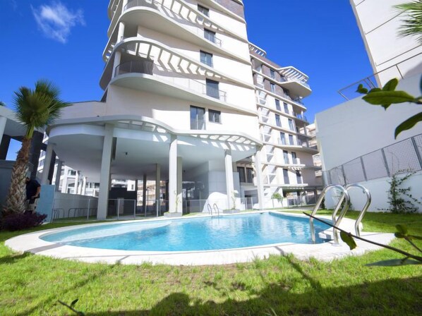 A modern multi-story building with a swimming pool surrounded by grass and palm trees.