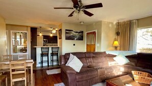 Ein Wohnzimmer mit braunem Ledersofa, einem Holztisch und einem Deckenventilator. Im Hintergrund ist eine Küche mit einer Bar und Stühlen zu sehen.