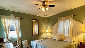 Ein Schlafzimmer mit einem Bett, einem Schreibtisch, einem Stuhl, einer Lampe, einem Deckenventilator und einem Fenster mit Vorhängen.