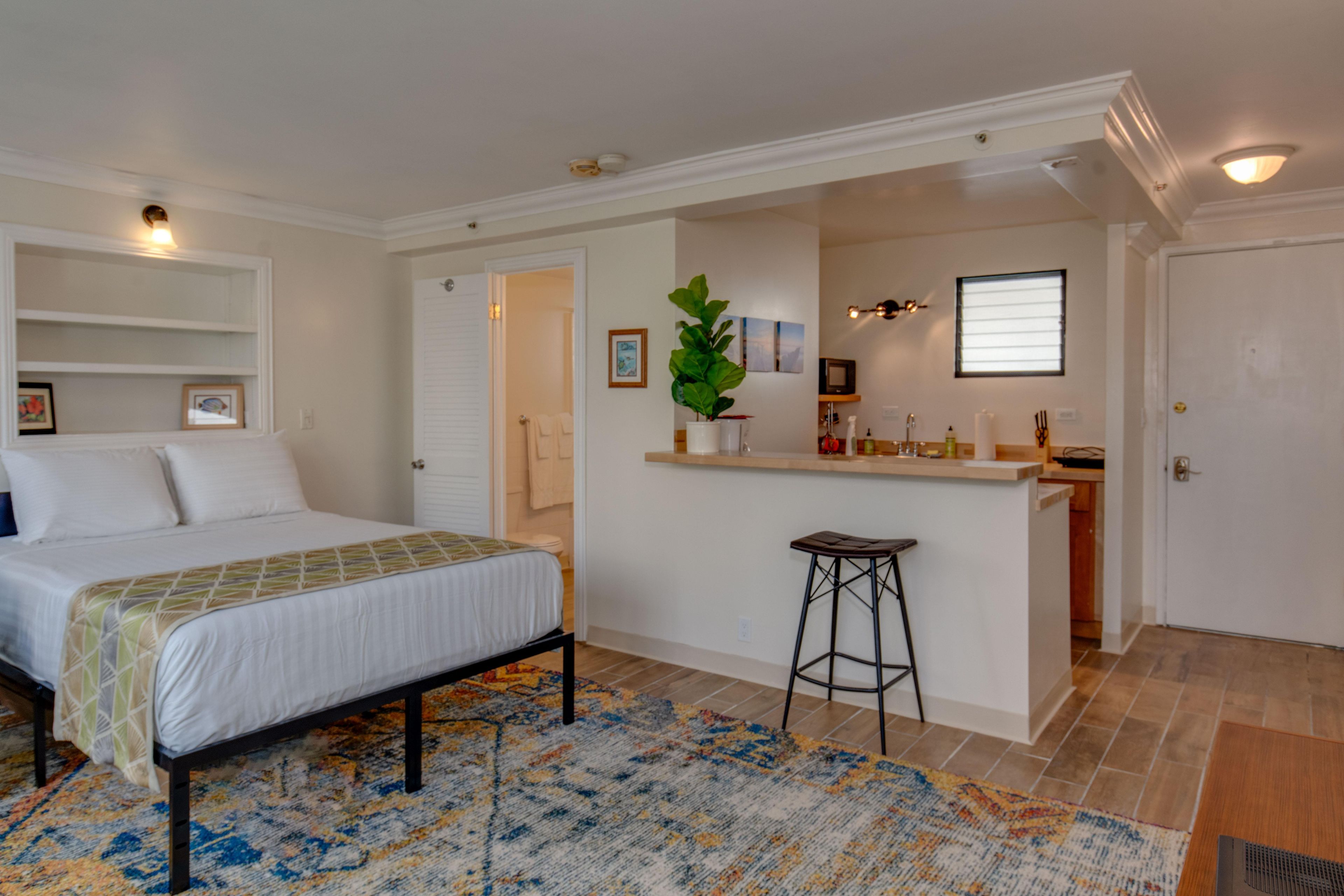 Open studio layout with a white bed, colorful patterned rug, and breakfast bar with a black stool.
