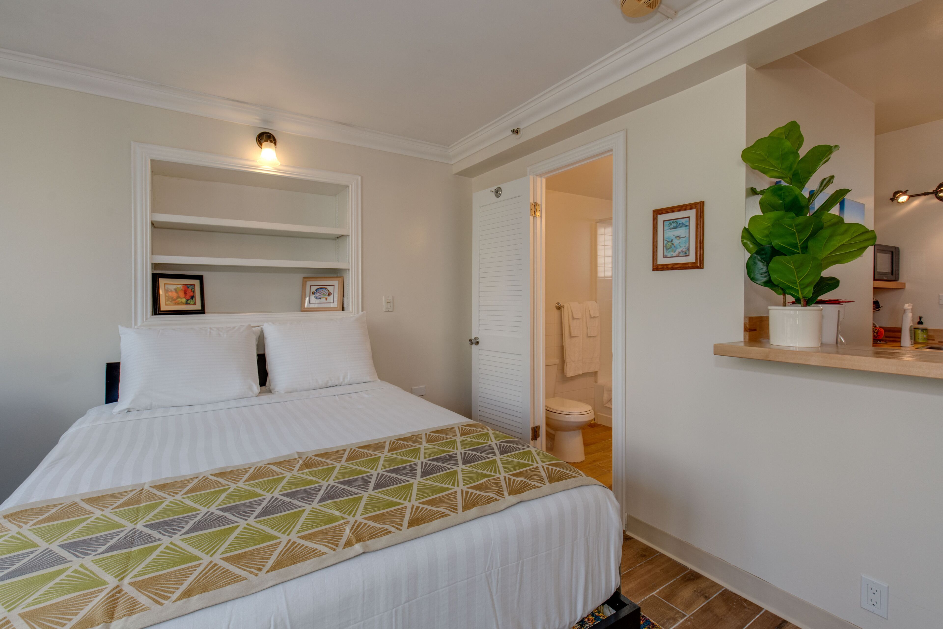 A neatly arranged bedroom with a bed, a bedside shelf, a mirror, and a potted plant.