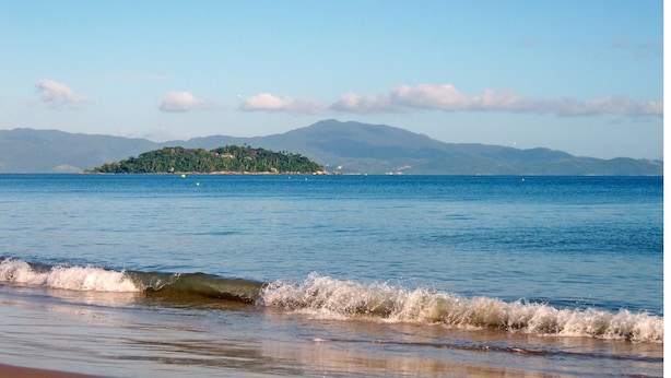 Una playa con olas, una isla distante y montañas al fondo.