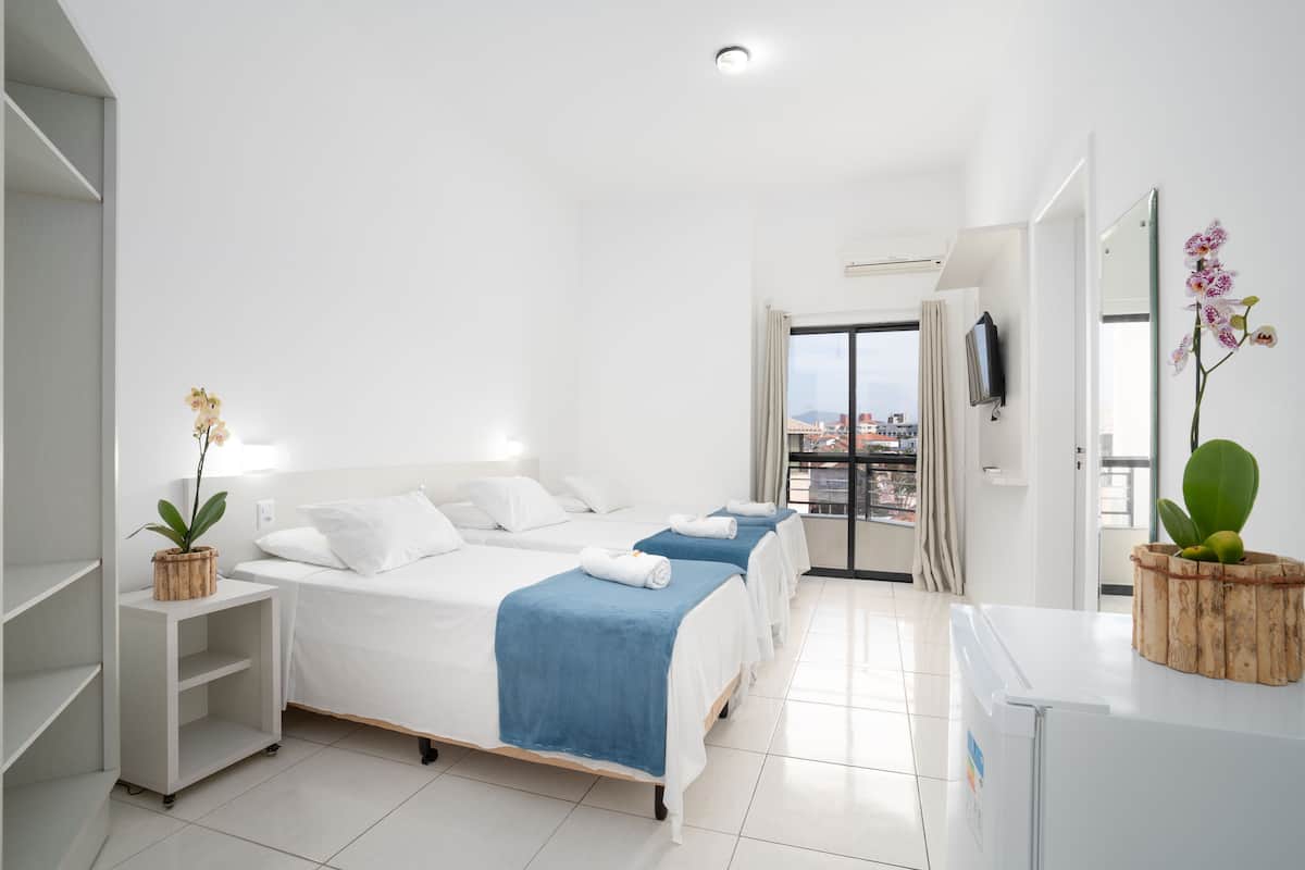 Une chambre d’hôtel avec deux lits, une armoire blanche, une petite table avec une plante, et une vue sur la ville par la fenêtre.