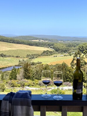 A scenic view from a balcony with two wine glasses and a bottle of wine, overlooking a lush valley with a lake and distant hills.