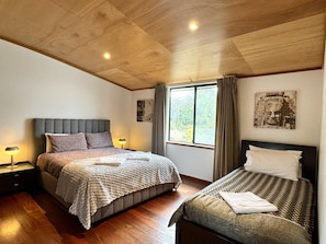 A bedroom with two beds, wooden flooring, a large window with curtains, and a ceiling with recessed lighting.