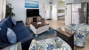 A living room with a sofa, coffee table, and a TV. There is a plant, a painting, and a refrigerator in the kitchen area.