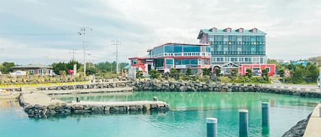 Un hotel frente al mar con un edificio grande de varios pisos y balcones, piscina y un litoral rocoso.