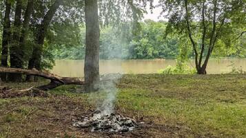 A natural outdoor setting with a campfire, trees, and a body of water in the background.