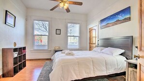 Een slaapkamer met een groot bed, een plafondventilator, twee ramen, een houten nachtkastje, een boekenkast en een schilderij aan de muur.