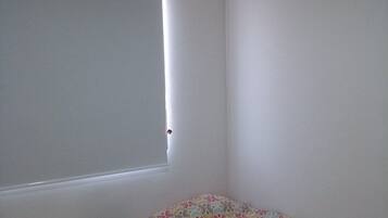 Een kleine, eenvoudig ingerichte kamer met twee bedden, een raam met zonwering en een plafondlamp.