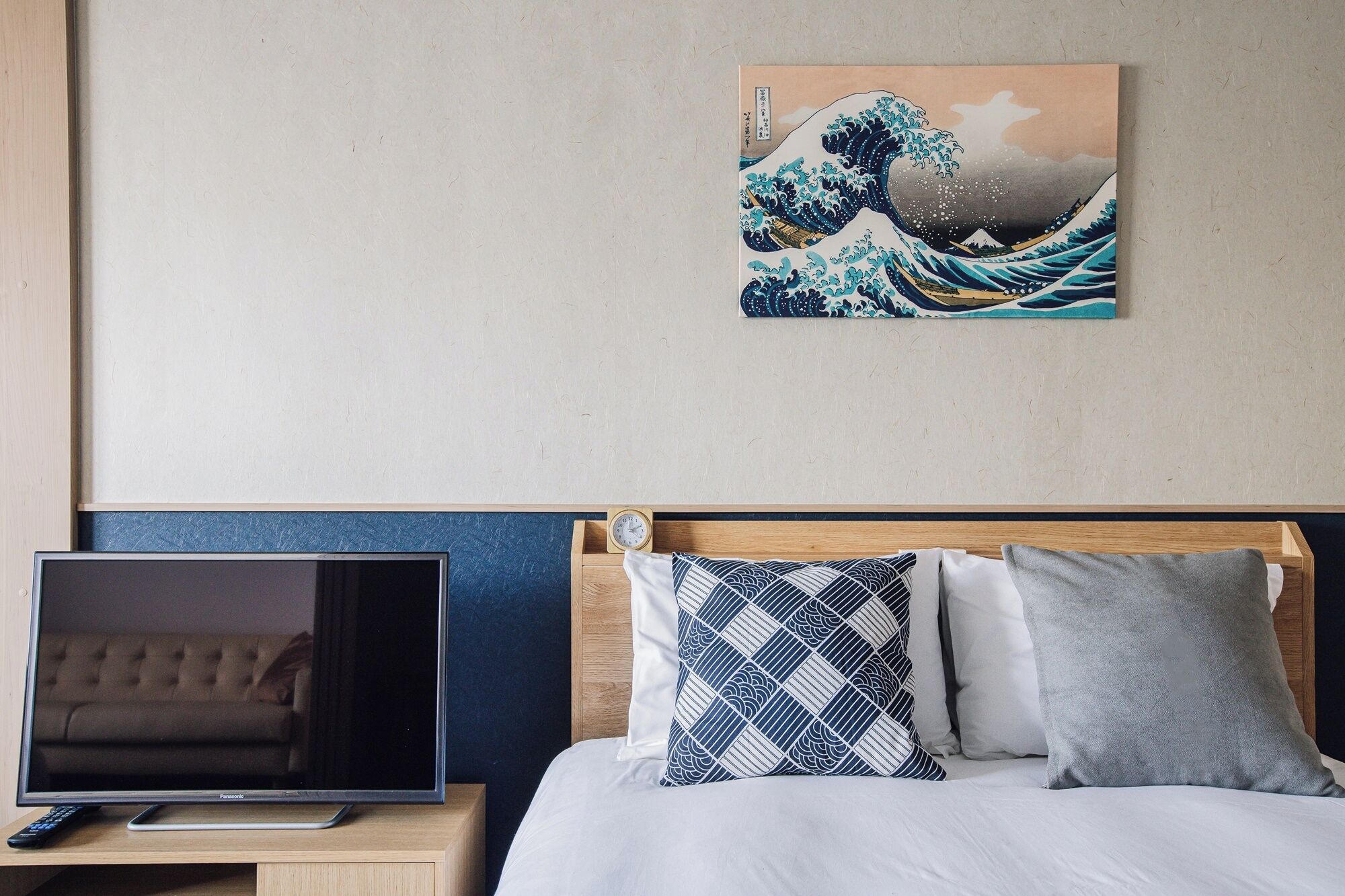 Habitación de hotel con cama, televisor sobre un soporte de madera y una pintura de una ola japonesa en la pared.