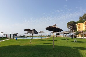 A pool area with thatched umbrellas, a fenced pool, and a building in the background.