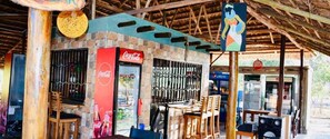 An outdoor cafe with a Coca-Cola and Pepsi refrigerator, wooden tables and chairs, and a thatched roof structure. - Ritungu Camp (Morogoro)