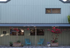 A building with a corrugated metal exterior, two windows with blinds, and a sign that reads "New Street Inn." There are two blue chairs outside and potted plants with flowers.