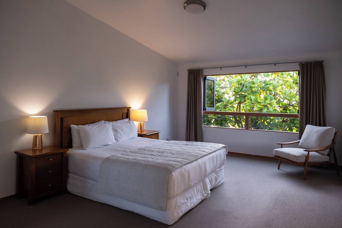 Habitación de hotel con una cama grande, dos lámparas de noche, una silla y una ventana con vistas a zonas verdes.
