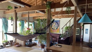 A spacious room with a wooden floor, a hammock, a tatami mat, a wooden table, and various potted plants. - Galaxy Guest House (Hachijo)