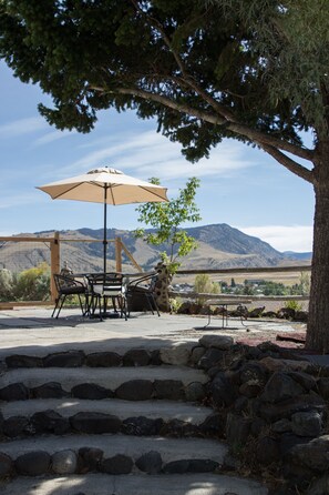 Patio esterno con tavolo e sedie, riparato da un ampio ombrellone, con vista panoramica su una montagna.