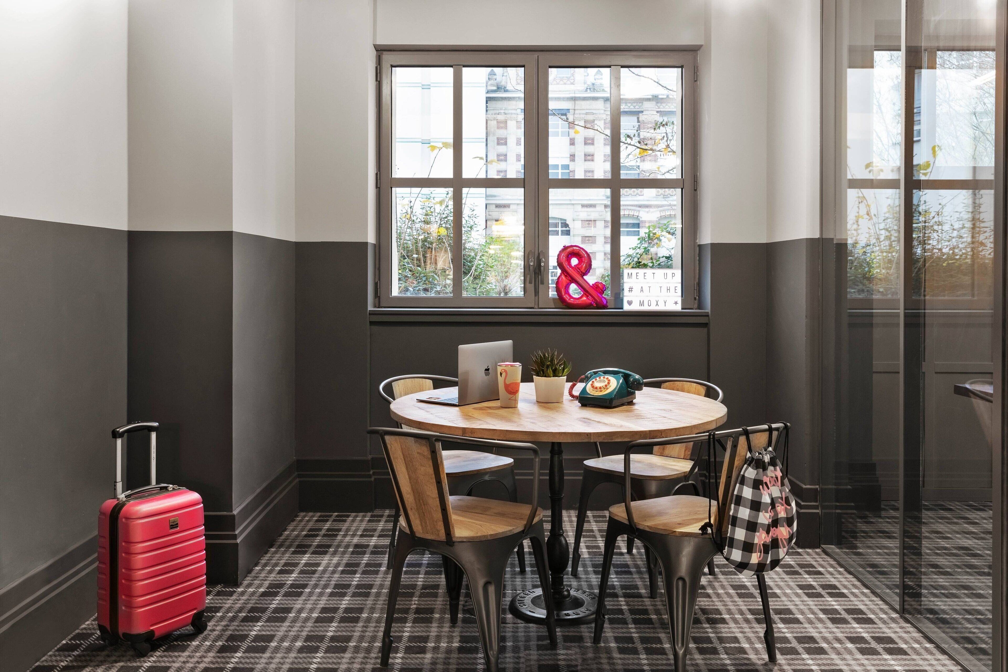 Une pièce avec une table ronde, des chaises, une fenêtre donnant sur un paysage, une valise rouge et un panneau lumineux au néon.