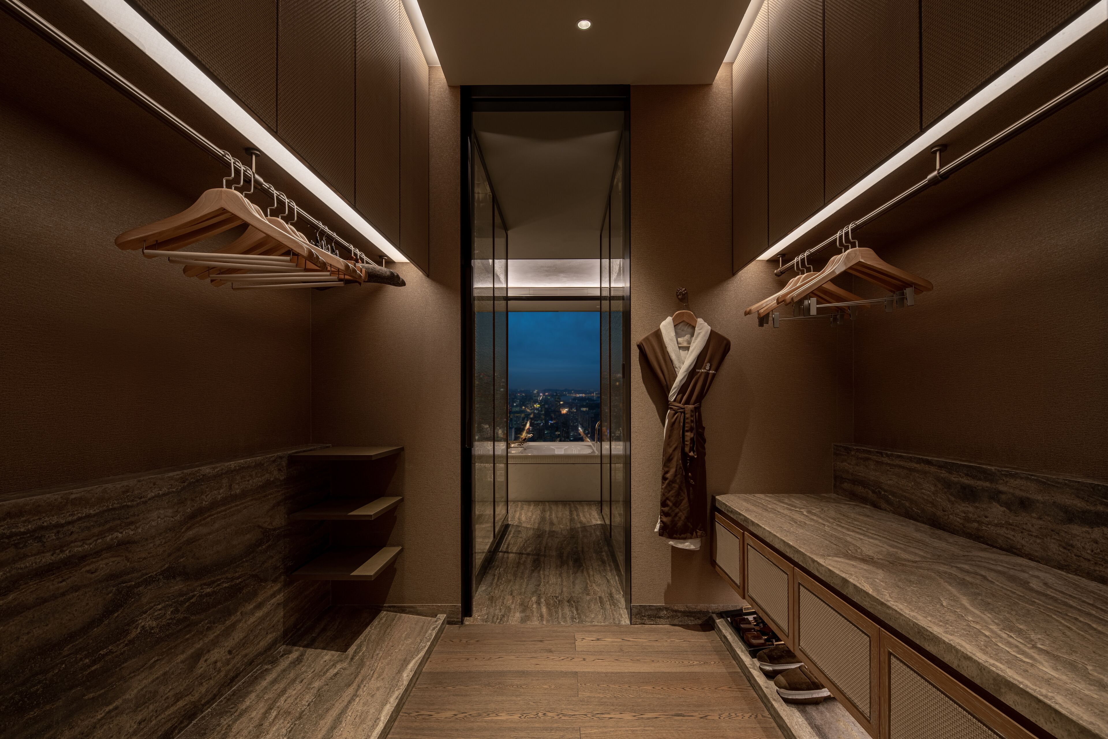 A modern hotel bathroom with wooden floors, marble countertops, and a view of the city at night.