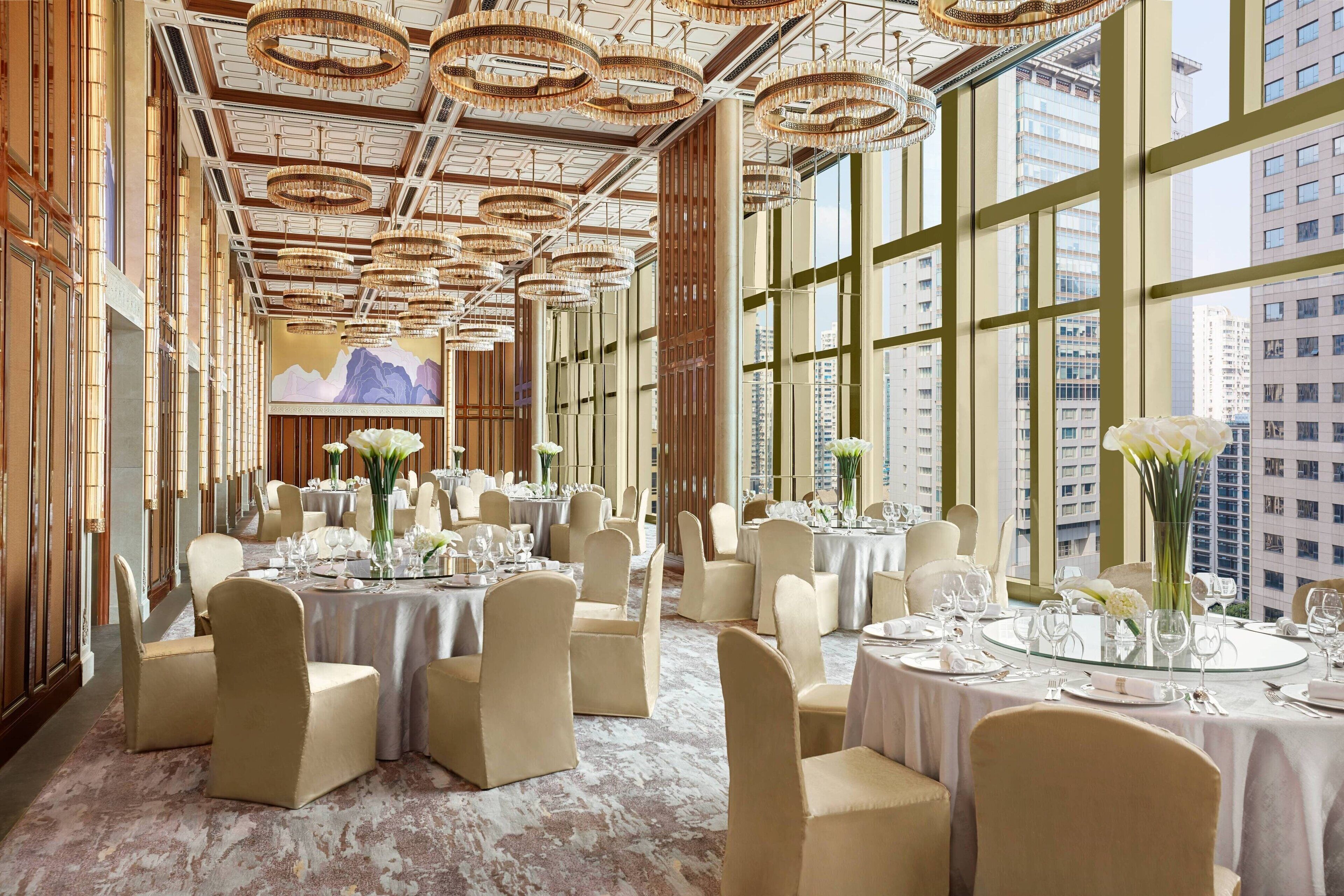 A spacious banquet hall with high ceilings, large windows, and elegant chandeliers. Tables are set for dining with chairs and centerpieces.