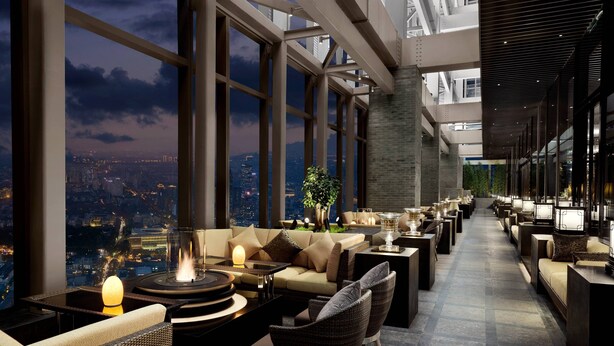 Un espace lounge moderne avec vue sur la ville, comprenant des canapés, des tables et un éclairage d’ambiance.