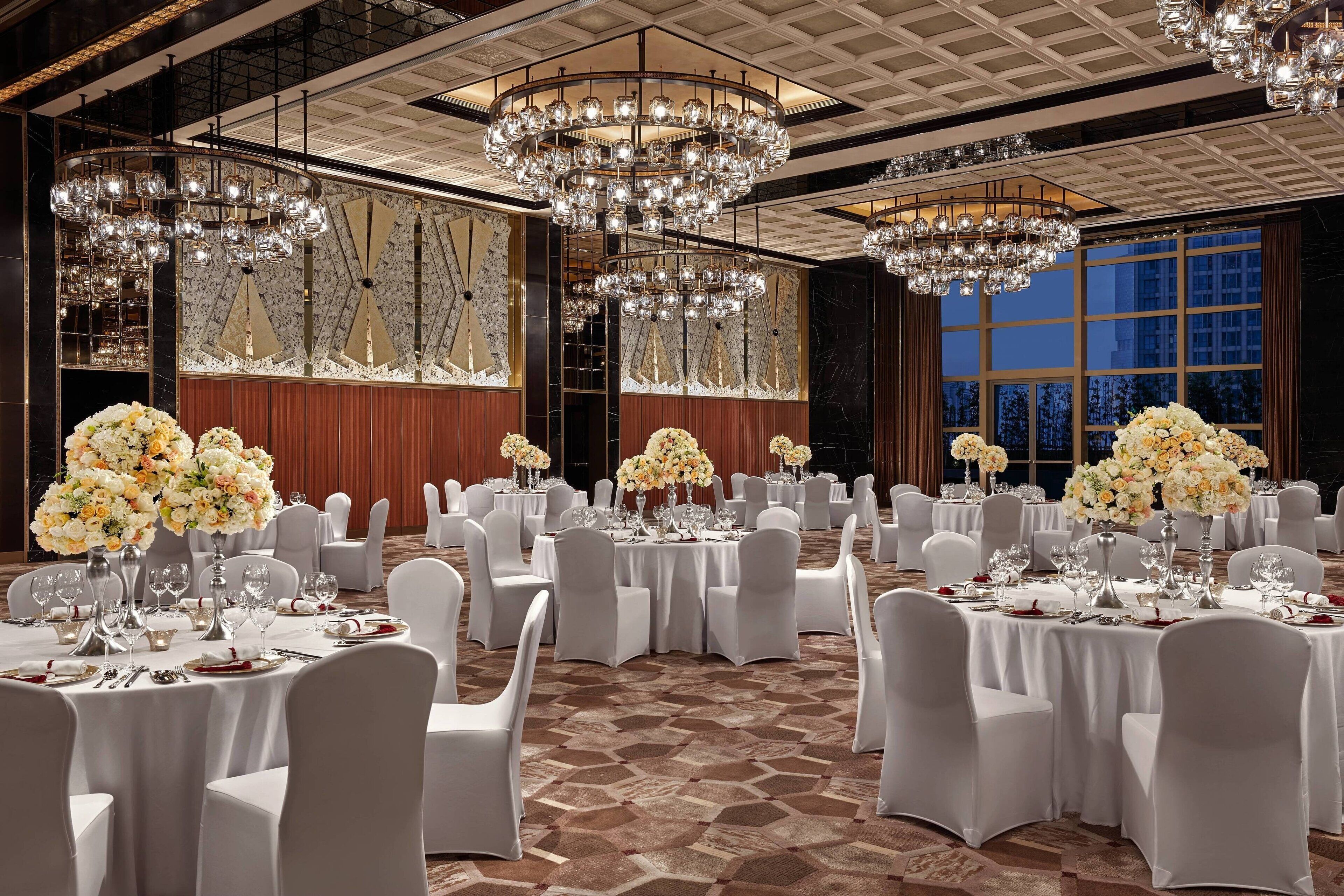 A banquet hall with round tables, white chairs, and chandeliers.