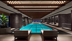 Une piscine intérieure moderne, dotée de chaises longues, d’un plafond en verre permettant de profiter de la lumière naturelle et offrant une vue sur le paysage urbain.