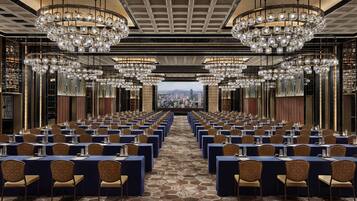 A conference room with rows of tables and chairs, large chandeliers, and a city view.