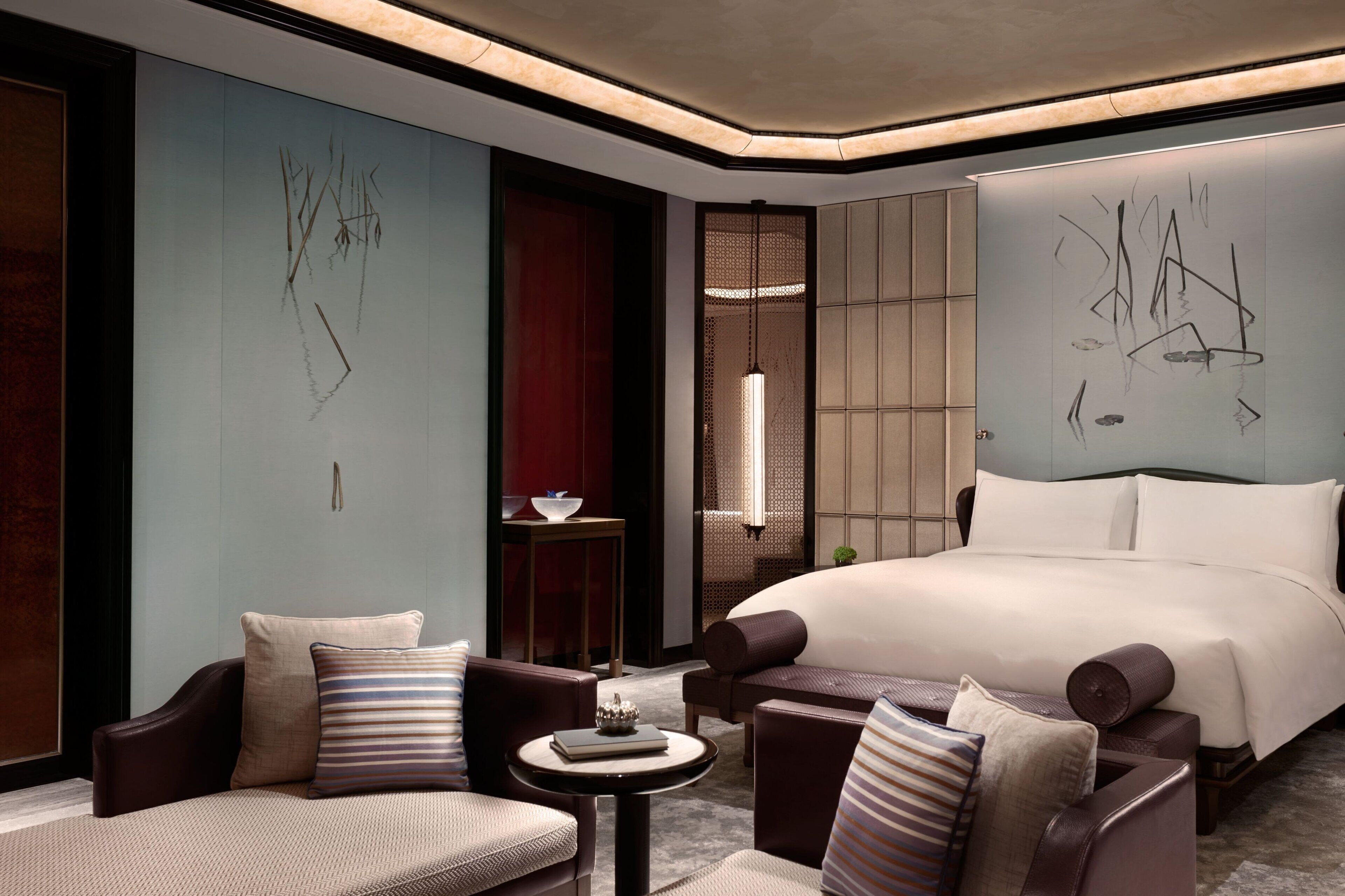 A modern hotel room with a large bed, two armchairs, a small table, and abstract wall art.
