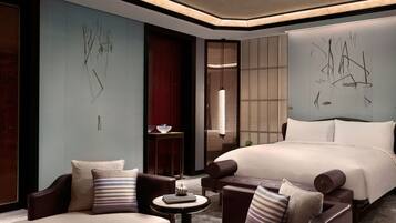 A modern hotel room with a large bed, two armchairs, a small table, and abstract wall art.