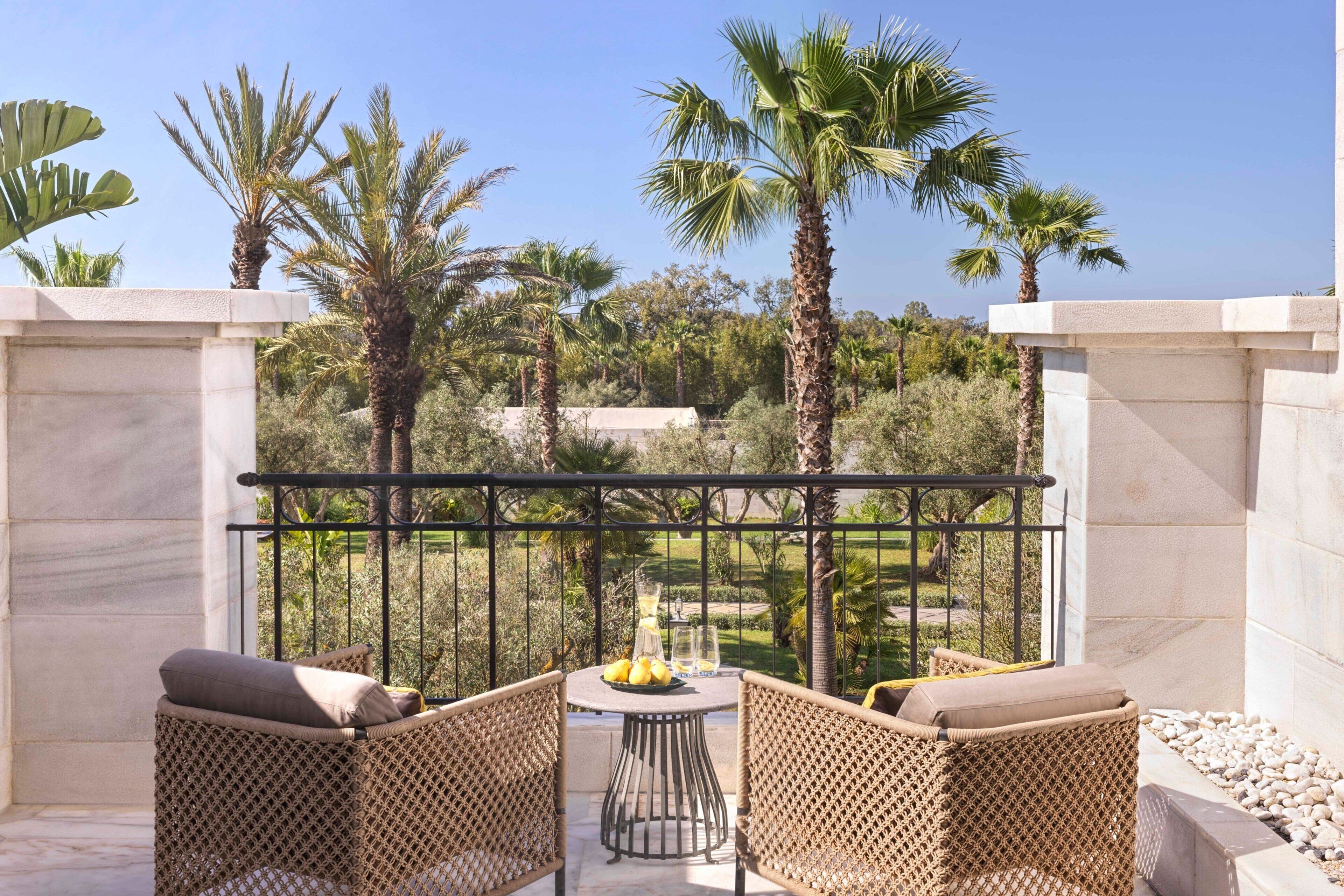 A balcony with wicker furniture, a table with a fruit bowl and glasses, and a view of palm trees and greenery.