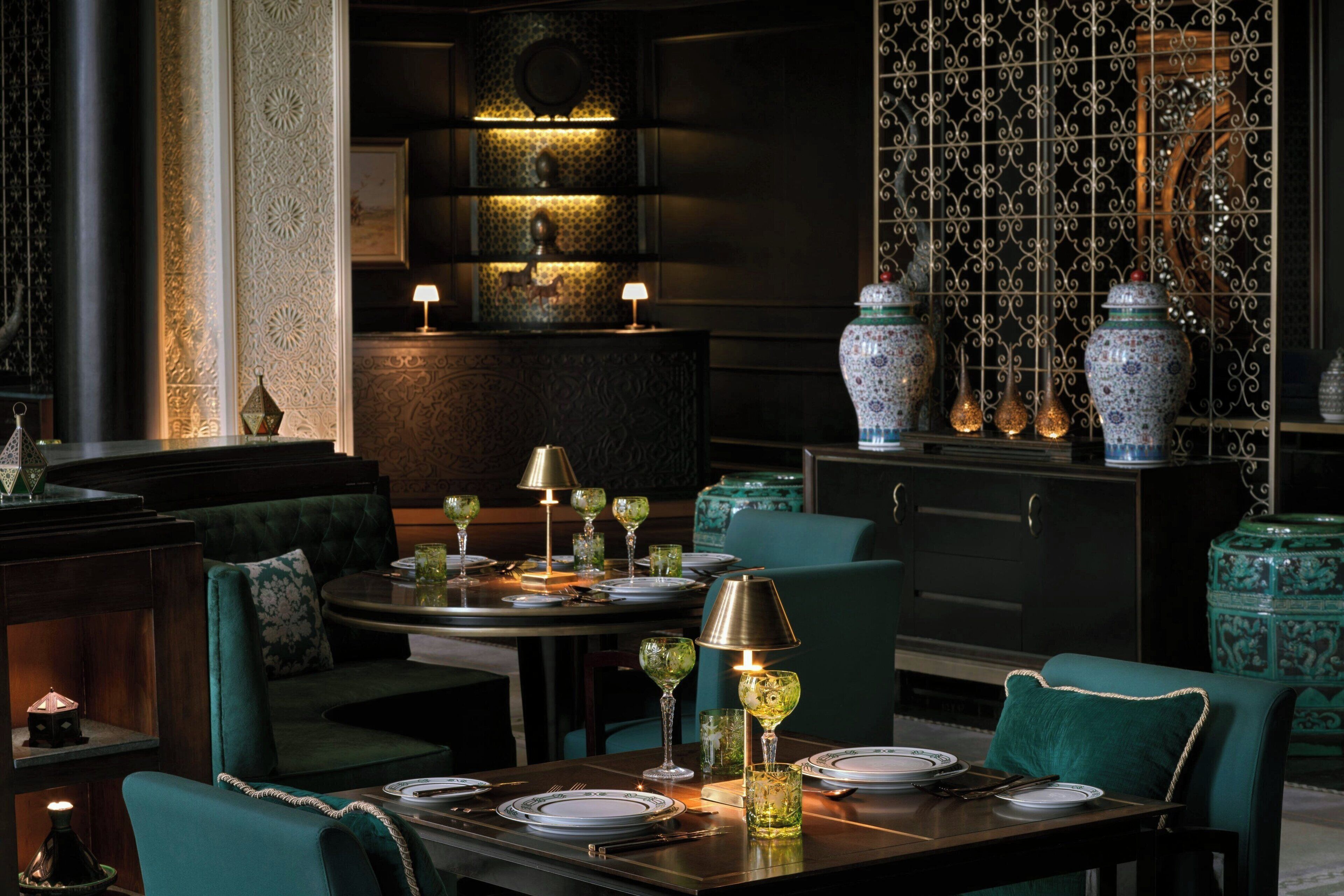 A dimly lit restaurant with a dark interior, green upholstered seating, and ornate decor including patterned wallpaper and traditional vases.