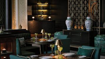 A dimly lit restaurant with a dark interior, green upholstered seating, and ornate decor including patterned wallpaper and traditional vases.