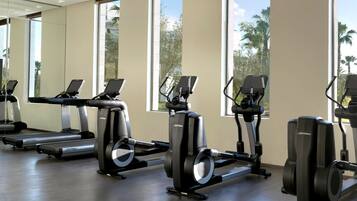 Un gimnasio con bicicletas estáticas y cintas de correr, ventanas amplias y vista a palmeras.