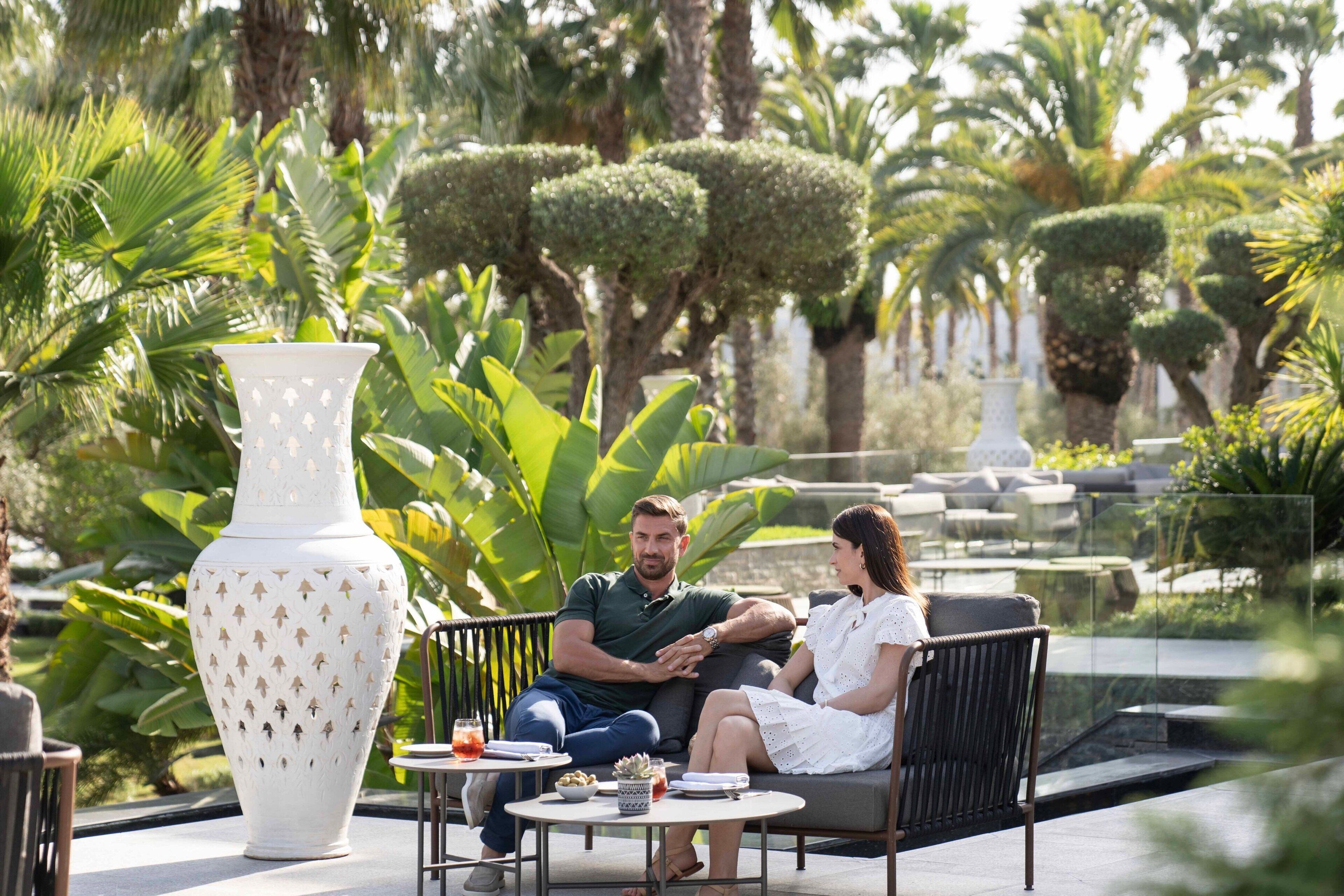 A couple seated on a bench in a garden with a large white vase and a small table with drinks.