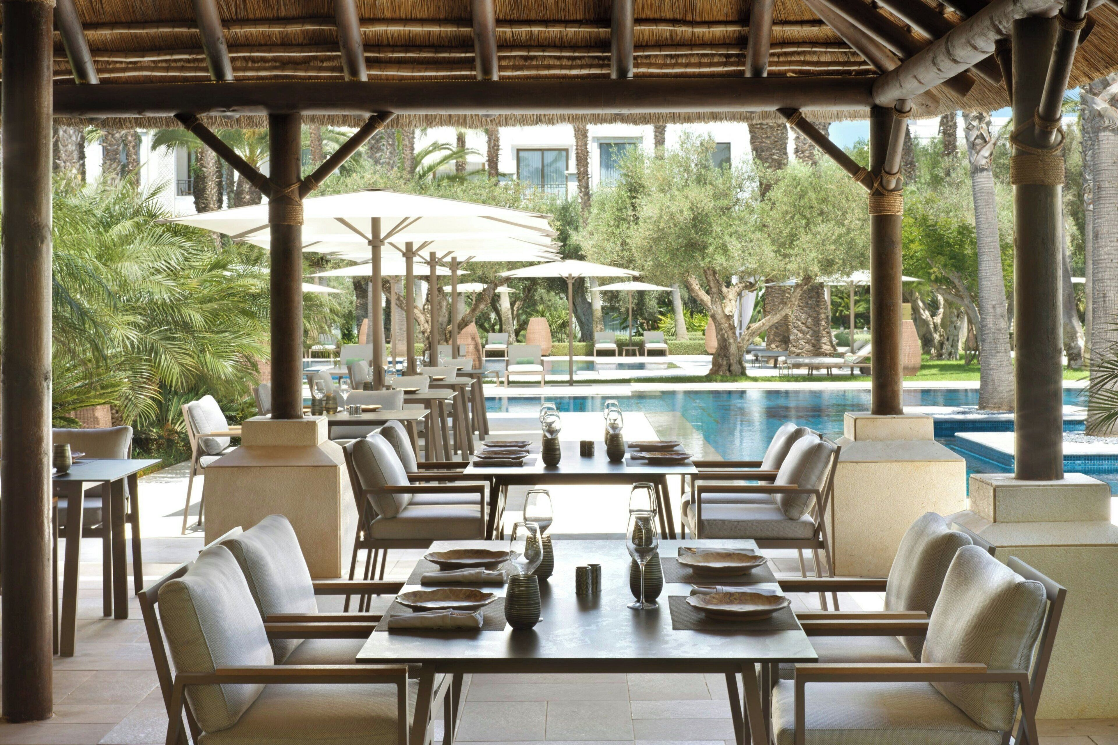 Un área de comedor al aire libre con mesas y sillas, una piscina al fondo y palmeras.