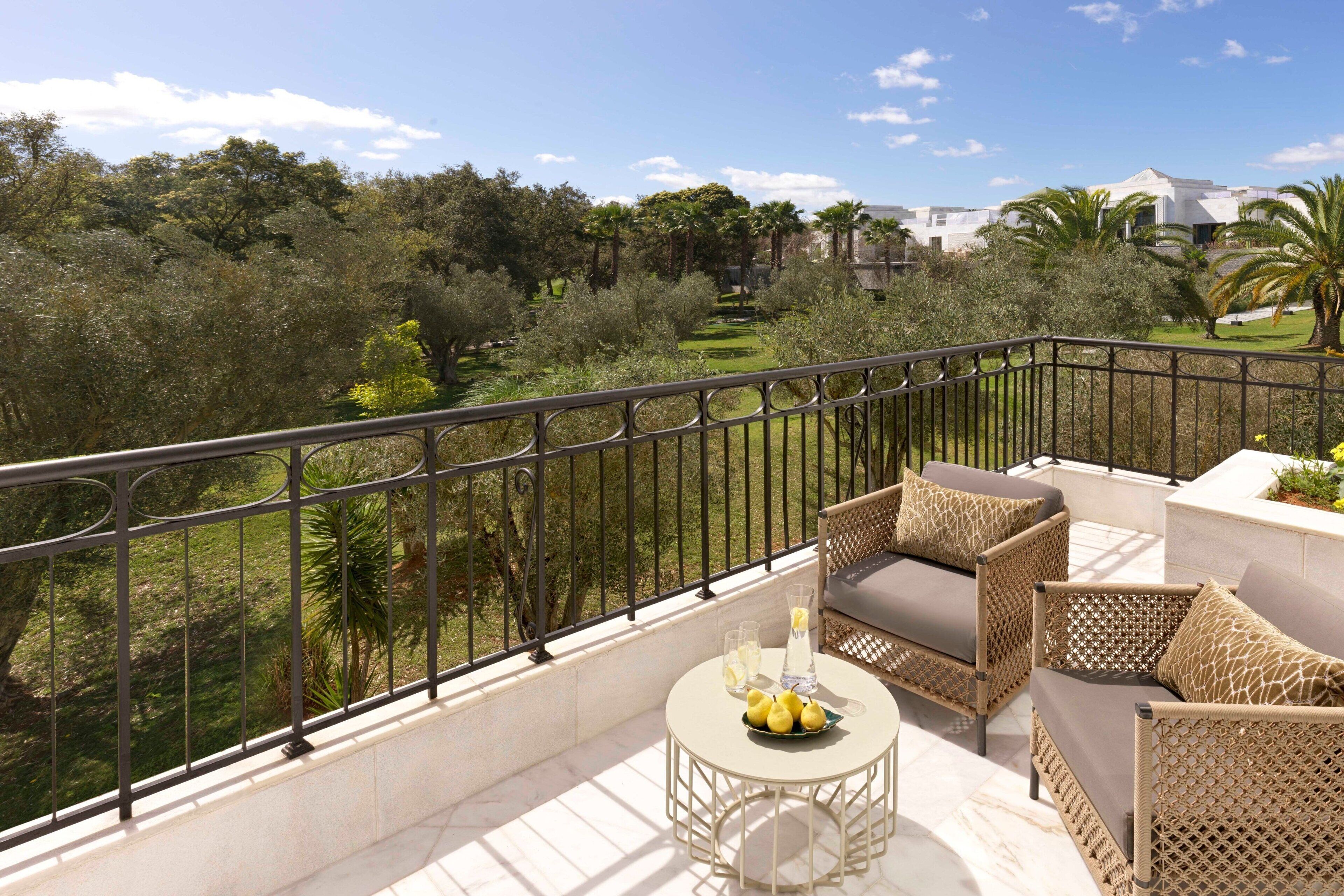 A balcony with wicker furniture, a small table with lemons, and a view of a lush garden and buildings in the background.