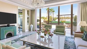 A spacious living room with a large flat-screen TV, a coffee table with floral centerpieces, a fireplace with intricate tile work, and a view of palm trees and a pool through large windows. - The Ritz-Carlton Rabat, Dar Es Salam (Rabat)
