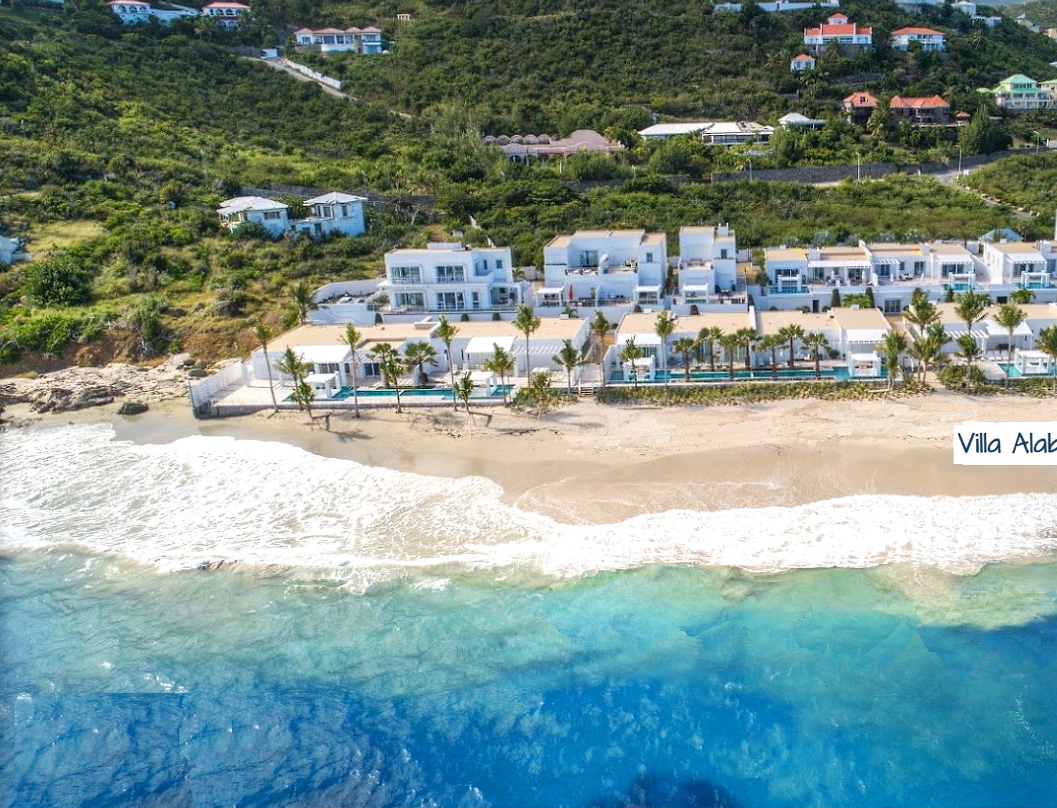 An aerial view of Villa Alabaster, featuring a beachfront property with multiple white buildings, a sandy beach, and a clear blue ocean.