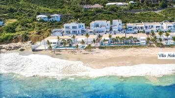 An aerial view of Villa Alabaster, featuring a beachfront property with multiple white buildings, a sandy beach, and a clear blue ocean.