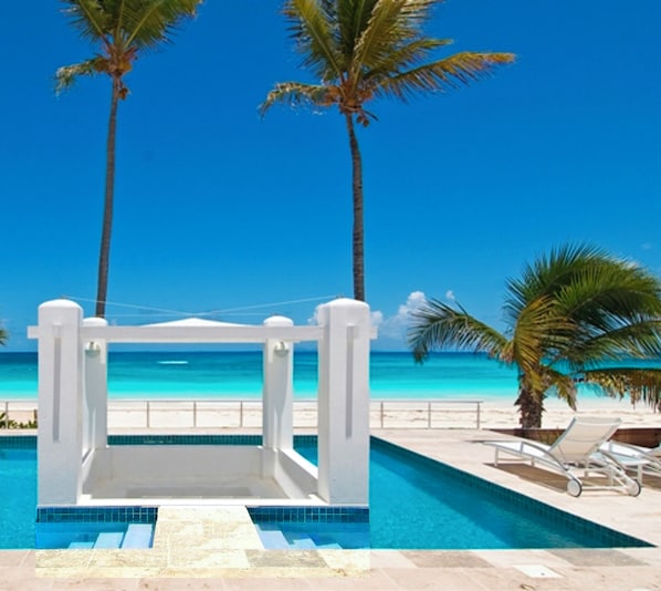 Zona de piscina con vista a la playa, palmeras y un cielo azul despejado.