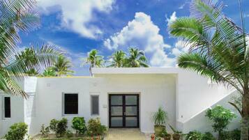 A white villa with a paved entrance, surrounded by lush greenery and palm trees.