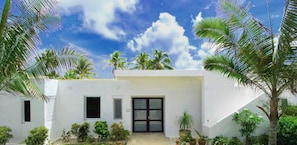 Una villa blanca con acceso pavimentado, rodeada de exuberante vegetación y palmeras.