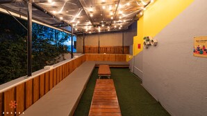 Un área de estar interior con bancos de madera y suelo verde, bajo un techo con vigas a la vista y luces colgantes.