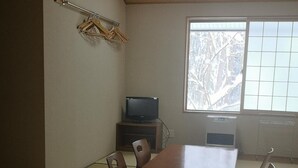 Una habitación con suelo de esteras de tatami, una mesa de madera y sillas. Un televisor está sobre un mueble bajo, y una ventana ofrece vistas a árboles.