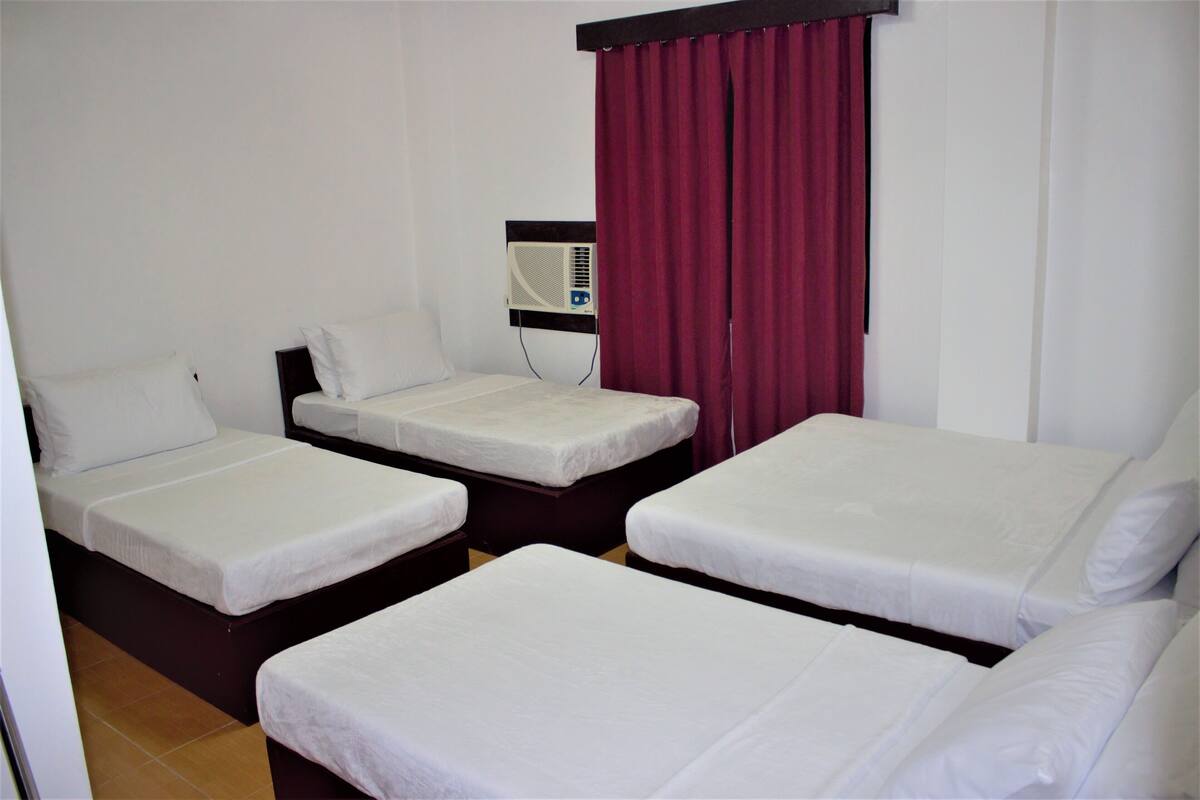 A hotel room with three single beds, a wall-mounted air conditioner, and a red curtain.