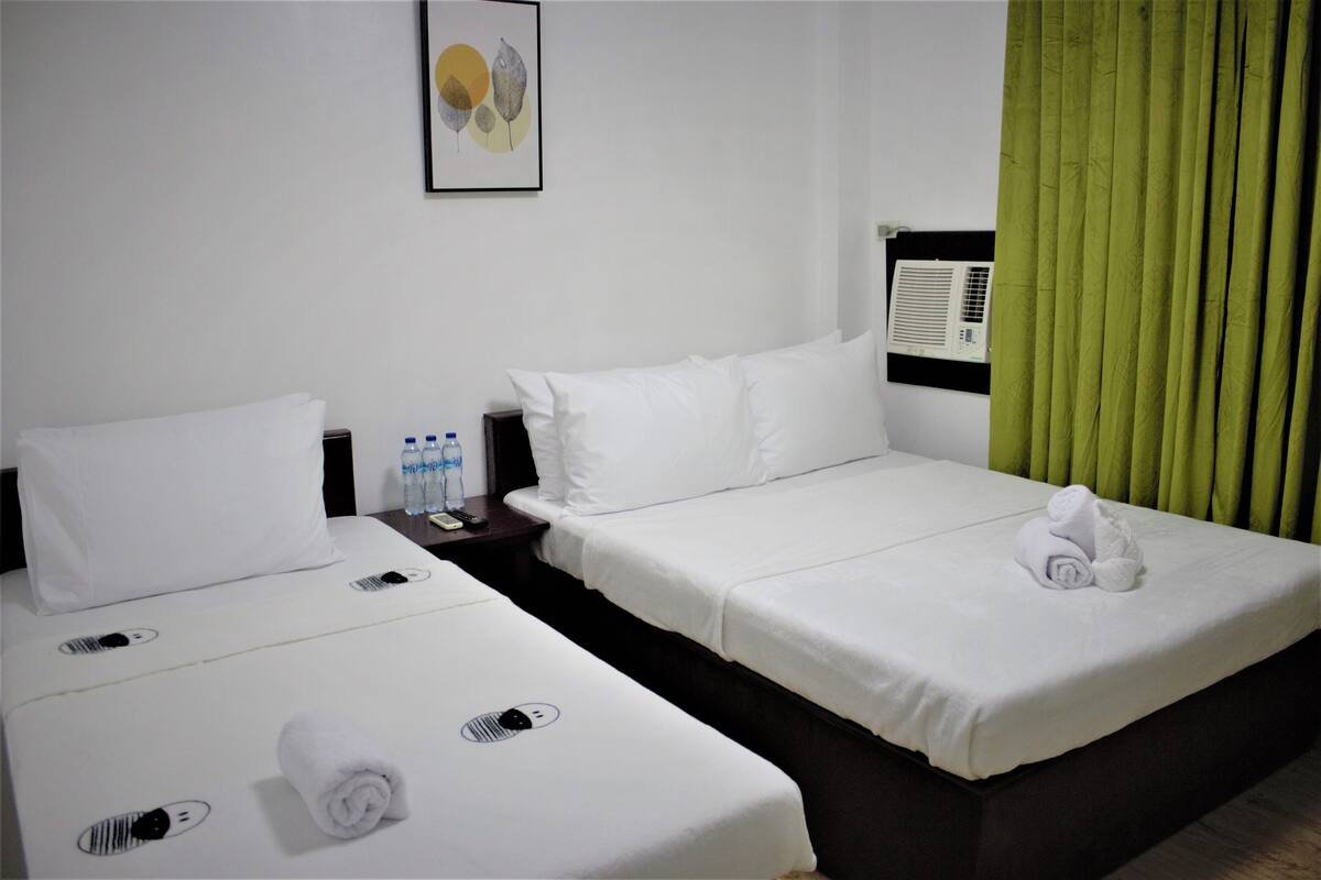 A hotel room with two beds, a nightstand with water bottles, a framed picture on the wall, and a green curtain.